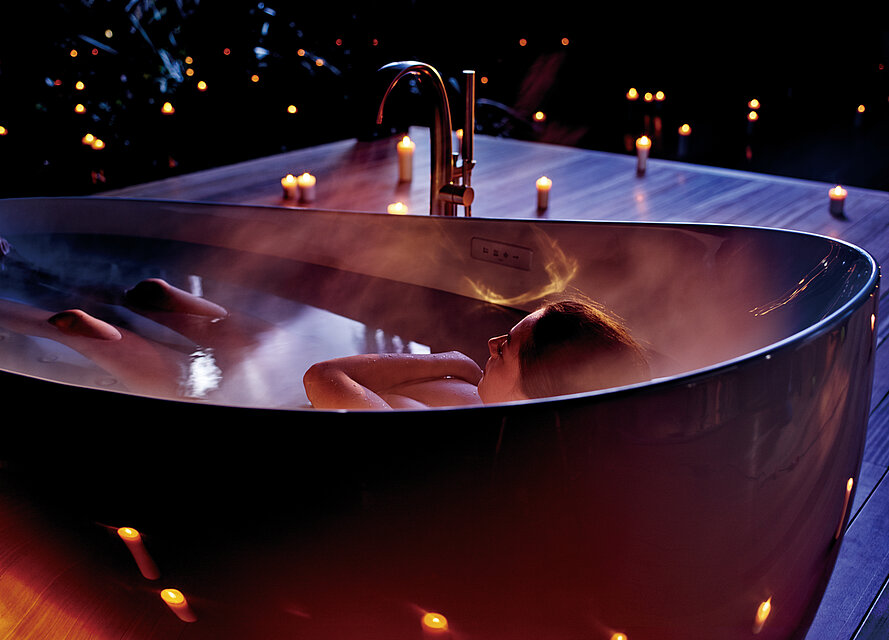 Eine Frau liegt in einer ovalen Badewanne. Alles außer ihren Knien, Kopf und über der Brust verschränkten Arme ist von Wasser bedeckt. Im Hintergrund stehen Kerzen.