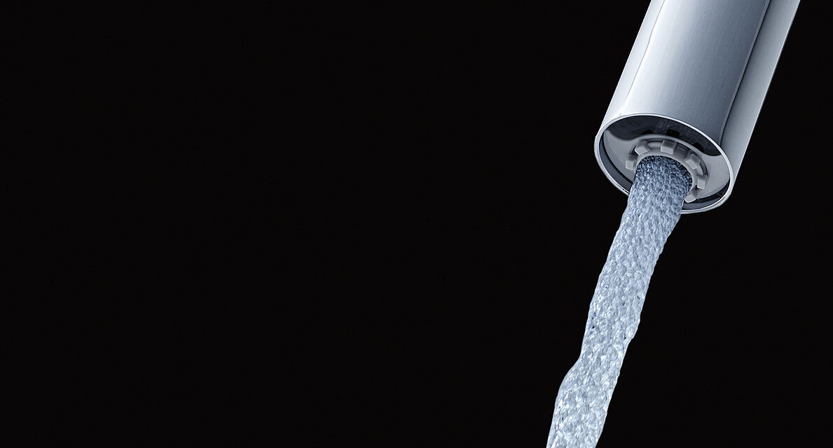 Detailaufnahme des Ausflusses eines Wasserhahns mit ECOCAP vor einem schwarzen Hintergrund. Der aus dem Hahn kommende Wasserstrahl ist mit kleinen Luftblasen durchsetzt. 