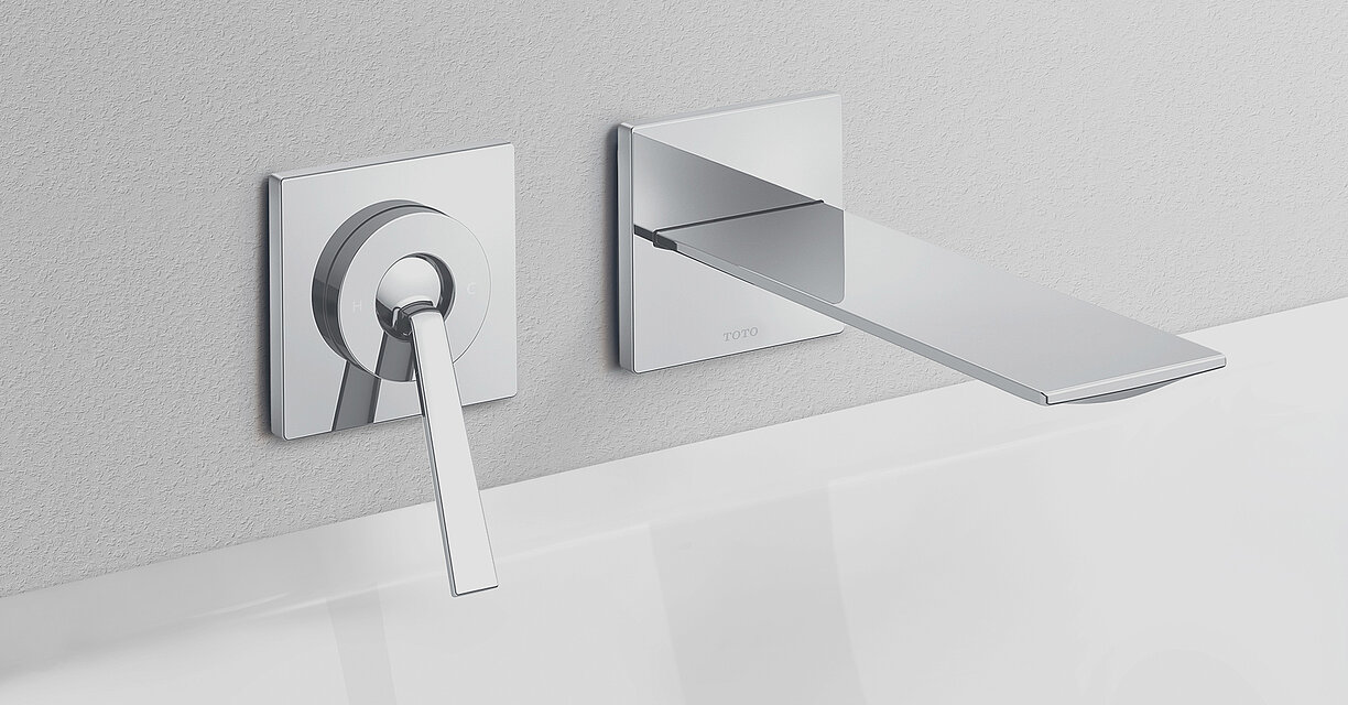 Auf dem Bild ist eine Armatur zu sehen. Sie besteht aus einem einzelnen, aus der Wand ragenden Arm und links daneben ist der Armaturen-hebel angebracht. Er befindet sich einige Zentimeter neben dem Wasserhahn. Beide Armaturen sind Chromfarben.