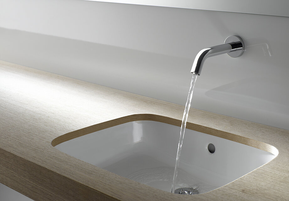 Eine Armatur ist an einer Wand über einem Waschtisch angebracht. Die Armatur ist eingeschaltet und aus ihr kommt ein Wasserstrahl. 