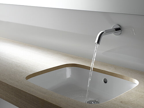 Eine Armatur ist an einer Wand über einem Waschtisch angebracht. Die Armatur ist eingeschaltet und aus ihr kommt ein Wasserstrahl. 
