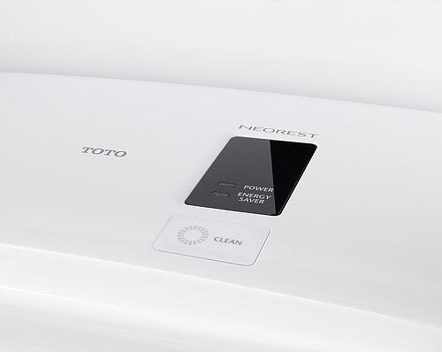 Man sieht eine Detailaufnahme der Anzeige auf der Oberseite des Washlet. In einem schwarzen Kasten stehen die Worte „Power“ und „Energy Saver“ neben zwei ausgeschalteten Lampen. Darüber befindet sich der Schriftzug „Neorest“, links daneben der Schriftzug TOTO und darunter in einem weißen Kasten der Schriftzug „Clean“ neben einem grauen Kreis.