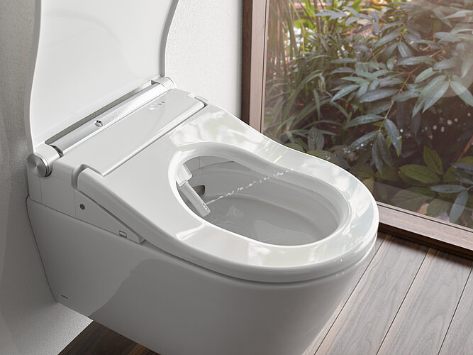 Das Bild zeigt eine Nahaufnahme eines modernen, wandhängenden TOTO WASHLETs mit geöffnetem Sitzdeckel und integrierter Bidetfunktion, sichtbar durch die im Sitz eingelassene Düse. Die Toilette steht neben einem Fenster, durch das man auf grüne Pflanzen blicken kann, was auf ein Badezimmer mit natürlichem Licht und Blick nach draußen hindeutet.