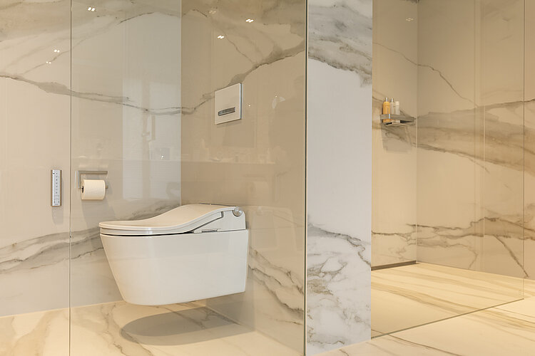 Das Bild zeigt ein Badezimmer mit einer modernen, wandhängenden Toilette vor einer Wand mit Marmorfliesen; daneben ist eine Glaswand, die vermutlich zu einer Duschkabine gehört. Auf der rechten Seite befindet sich ein eingebautes Wandregal mit Kerzen, und die gesamte Szene strahlt durch die harmonische Farbwahl und das Design eine ruhige und elegante Atmosphäre aus.