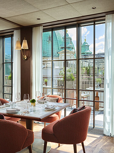 Das Bild zeigt ein ansprechend gedecktes Esszimmer mit einem runden Glas-Esstisch und rostfarbenen Stühlen, beleuchtet von eleganten Pendelleuchten. Durch die großen Fenster bietet sich ein Blick auf eine Terrasse und die historische Architektur im Hintergrund, was auf eine städtische Lage hindeutet.