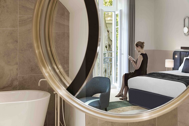 Das Foto zeigt einen Blick durch einen runden, an der Wand montierten Spiegel auf eine stilvolle Suite mit modernem Interieur. Durch den Spiegel hindurch ist eine Frau in einem eleganten Kleid sichtbar, die in einem Sessel am Balkon sitzt und ein Buch liest, während im Vordergrund des Spiegels eine freistehende Badewanne zu erkennen ist.
