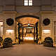 Das Bild zeigt den festlich beleuchteten Eingangsbereich eines eleganten Hotels, geschmückt mit Kränzen und Lichterketten. Laternen und mit Lichtern dekorierte Pflanzenkübel flankieren den Weg, der zu den automatischen Glastüren führt, und verleihen dem Eingang eine einladende und festliche Atmosphäre.