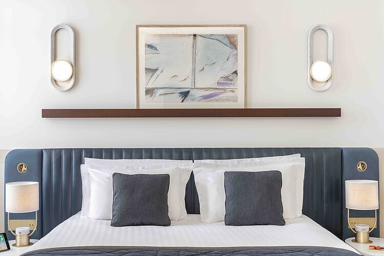 Das Bild zeigt das obere Ende eines Bettes in einem hell gestalteten Raum, mit einem dunkelblauen, getufteten Kopfteil, das mit mehreren Kissen in Grau und Weiß ausgestattet ist. Über dem Bett hängt ein gerahmtes Kunstwerk, flankiert von zwei modernen, ovalen Wandlampen, die ein warmes Licht abgeben.